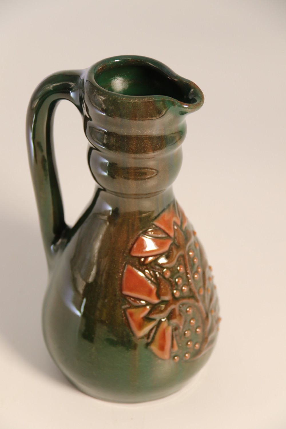 A green glazed ceramic jug featuring a flared spout, loop handle, and an embossed sunflower motif on the side. View X