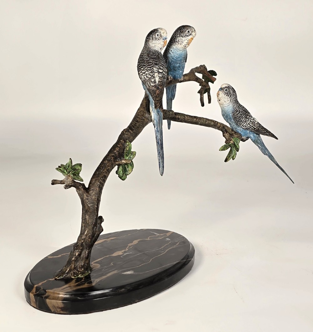A cold painted bronze sculpture depicting three blue budgerigars perched on a branch mounted on an oval dark marble base.