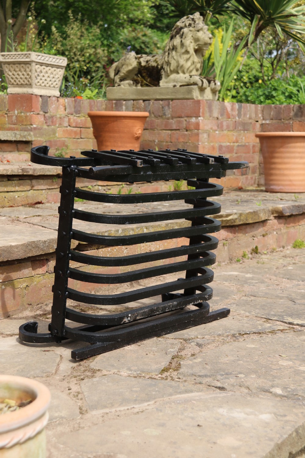 A black wrought iron fire basket featuring a curved slatted log cradle, scrolled corner terminals, and riveted front bars. View III