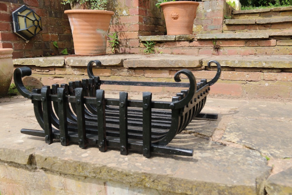 A black wrought iron fire basket featuring a curved slatted log cradle, scrolled corner terminals, and riveted front bars. View VII