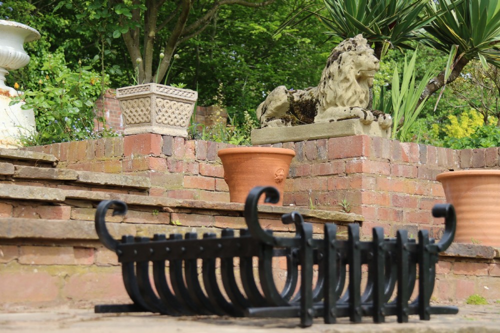 A black wrought iron fire basket featuring a curved slatted log cradle, scrolled corner terminals, and riveted front bars. View IX