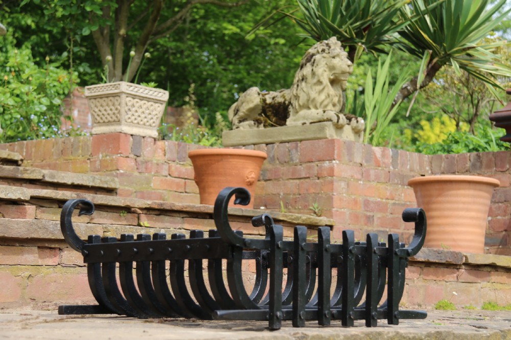 A black wrought iron fire basket featuring a curved slatted log cradle, scrolled corner terminals, and riveted front bars. View X