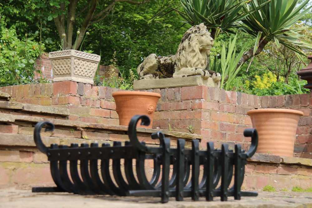 A black wrought iron fire basket featuring a curved slatted log cradle, scrolled corner terminals, and riveted front bars. View XI