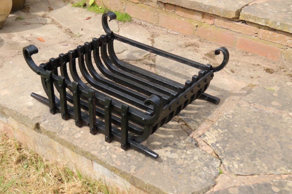 A black wrought iron fire basket featuring a curved slatted log cradle, scrolled corner terminals, and riveted front bars. View XV