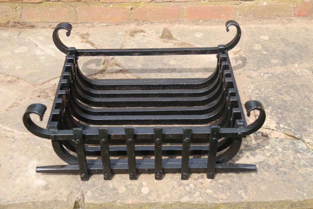 A black wrought iron fire basket featuring a curved slatted log cradle, scrolled corner terminals, and riveted front bars.