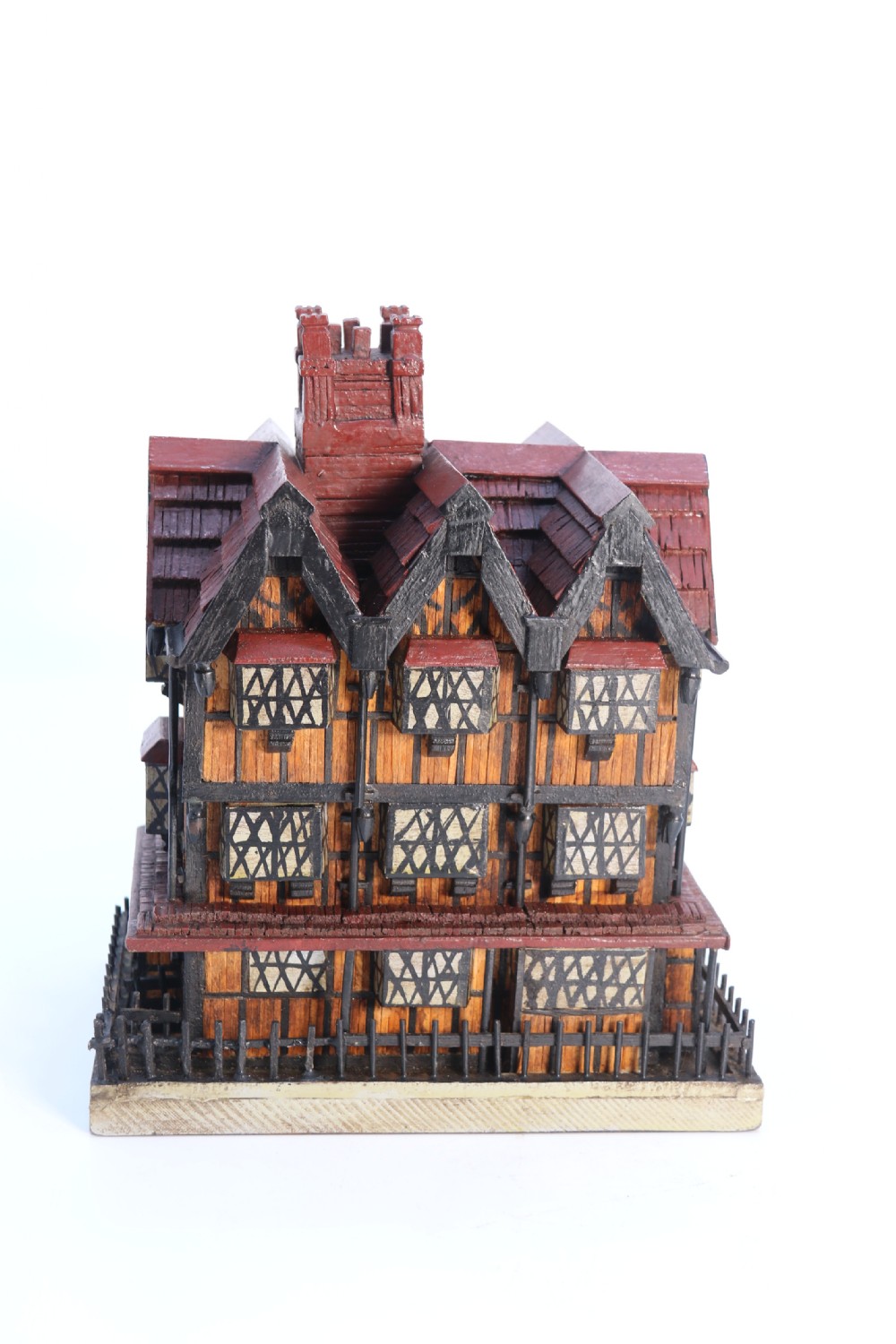 A hand-crafted matchstick model of a three-storey Tudor style half-timbered house featuring red gabled roof tiles, black timber details, and leaded window patterns on a rectangular wooden base. View III