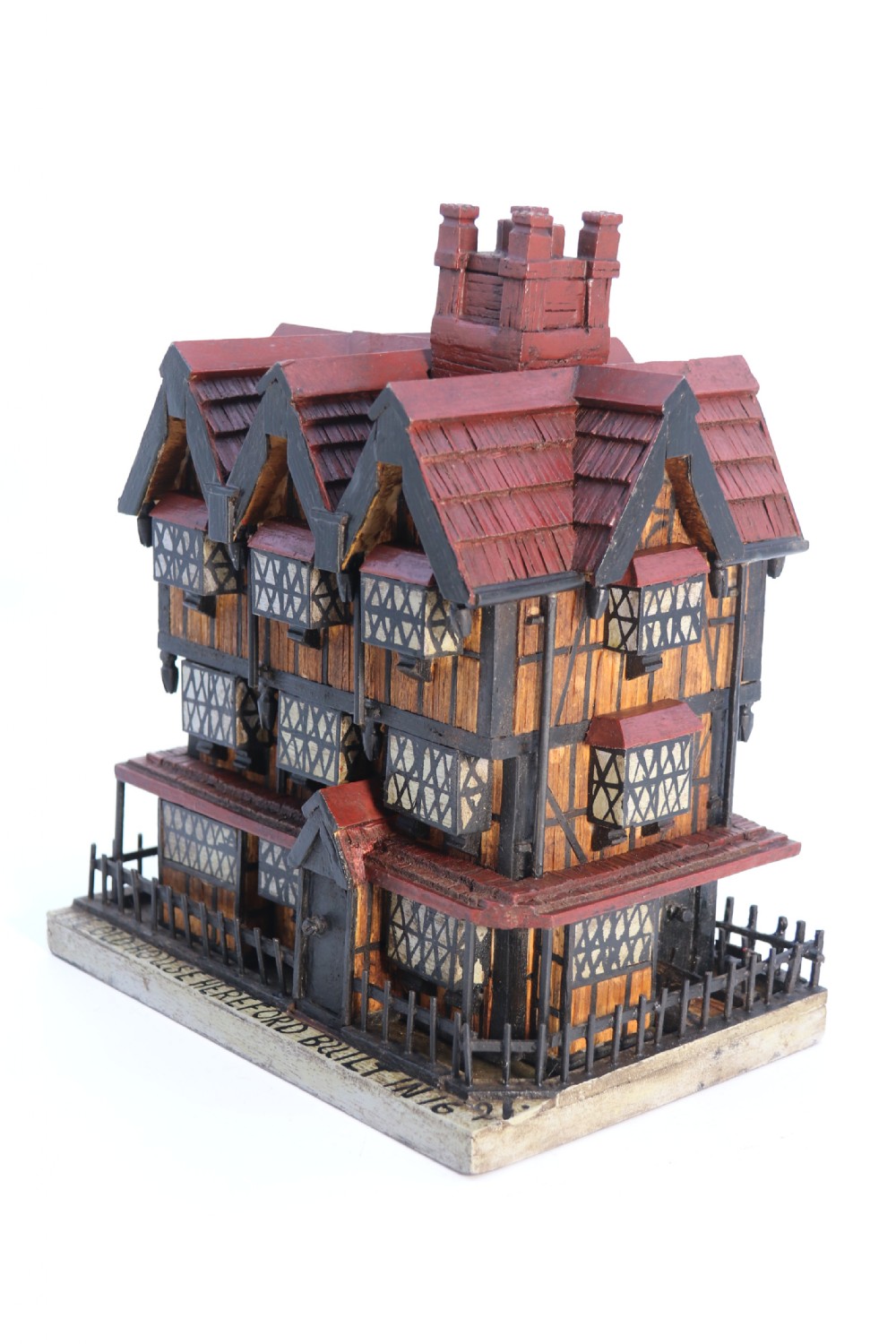 A hand-crafted matchstick model of a three-storey Tudor style half-timbered house featuring red gabled roof tiles, black timber details, and leaded window patterns on a rectangular wooden base. View VI