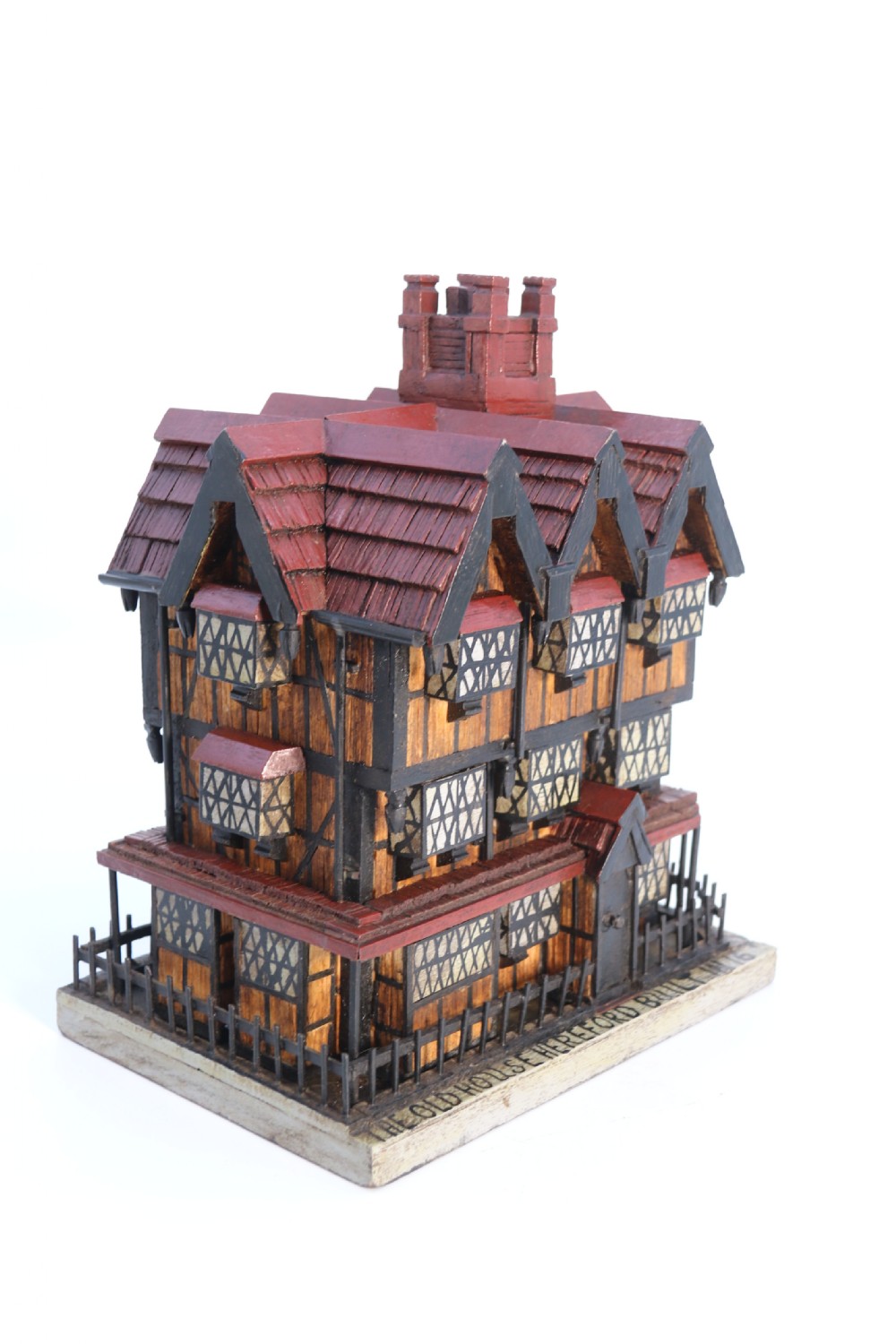 A hand-crafted matchstick model of a three-storey Tudor style half-timbered house featuring red gabled roof tiles, black timber details, and leaded window patterns on a rectangular wooden base. View VII