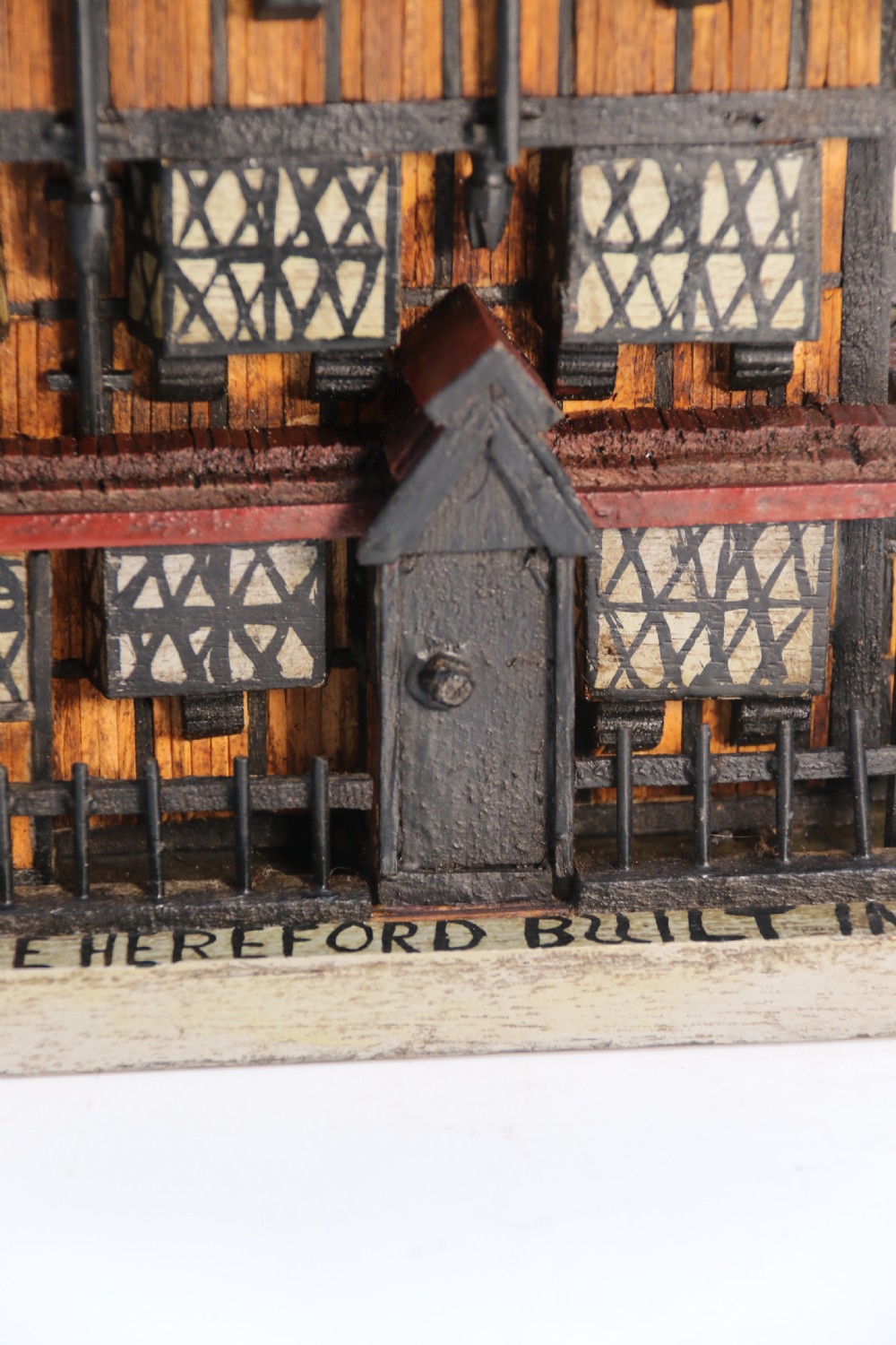 A hand-crafted matchstick model of a three-storey Tudor style half-timbered house featuring red gabled roof tiles, black timber details, and leaded window patterns on a rectangular wooden base. View IX
