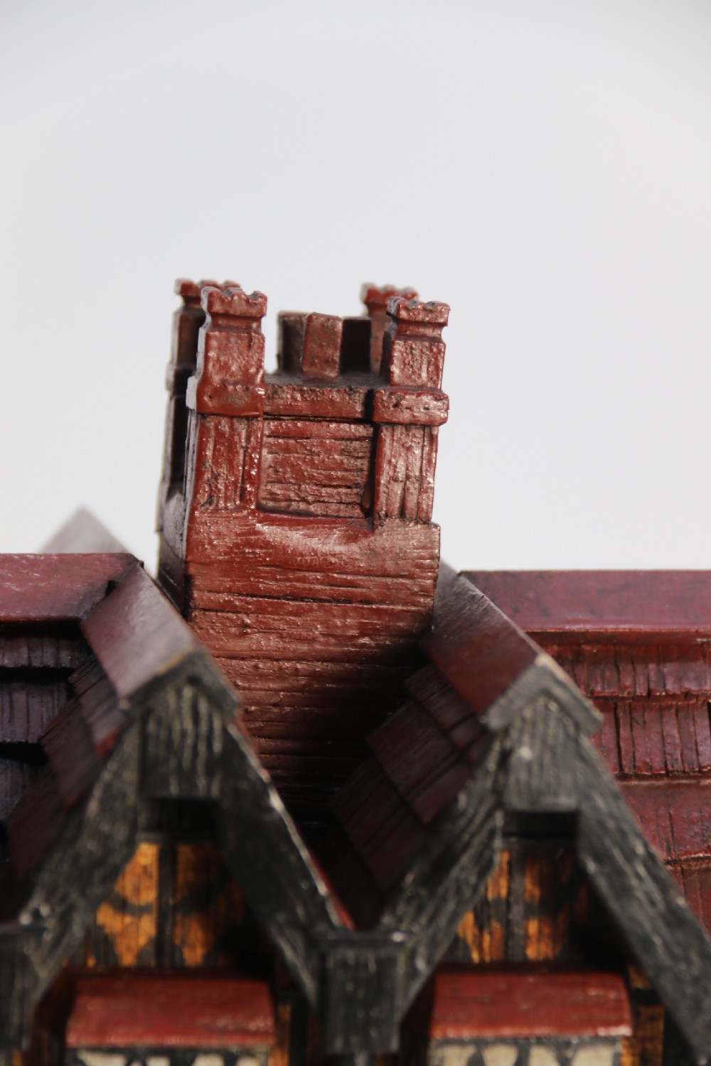 A hand-crafted matchstick model of a three-storey Tudor style half-timbered house featuring red gabled roof tiles, black timber details, and leaded window patterns on a rectangular wooden base. View X