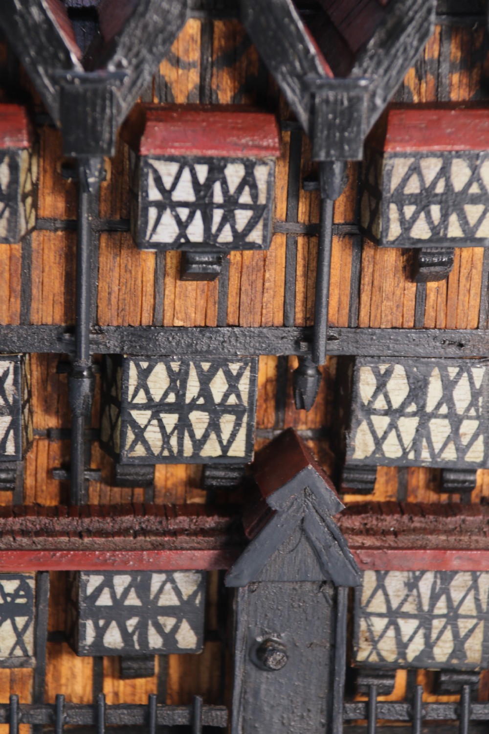A hand-crafted matchstick model of a three-storey Tudor style half-timbered house featuring red gabled roof tiles, black timber details, and leaded window patterns on a rectangular wooden base. View XI