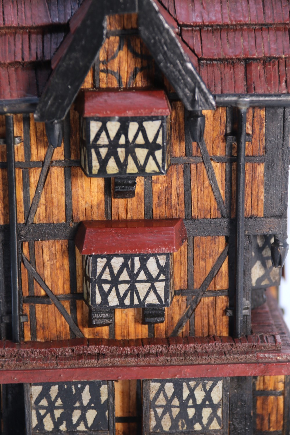 A hand-crafted matchstick model of a three-storey Tudor style half-timbered house featuring red gabled roof tiles, black timber details, and leaded window patterns on a rectangular wooden base. View XII