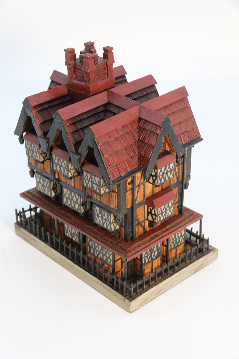A hand-crafted matchstick model of a three-storey Tudor style half-timbered house featuring red gabled roof tiles, black timber details, and leaded window patterns on a rectangular wooden base. View XVIII