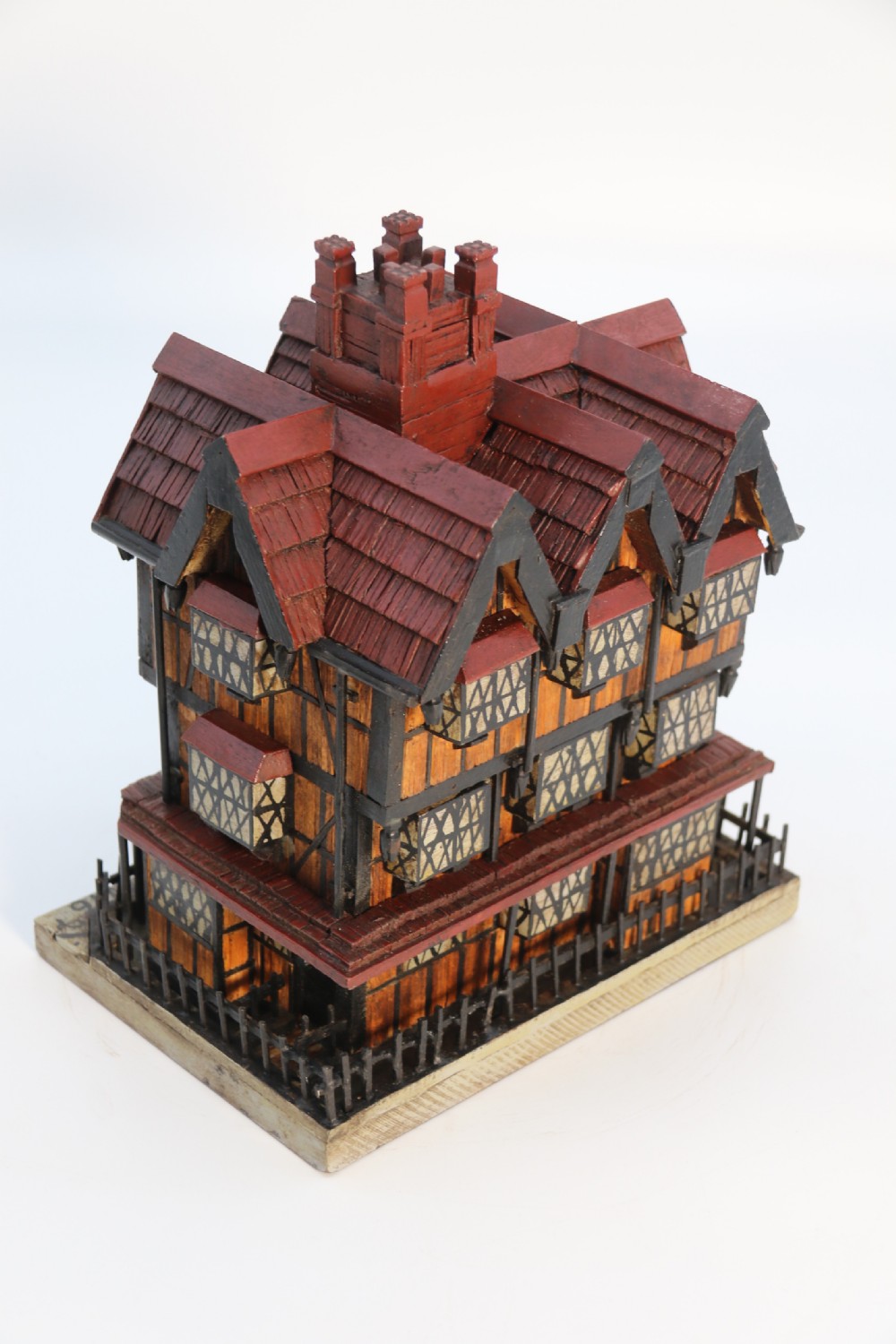 A hand-crafted matchstick model of a three-storey Tudor style half-timbered house featuring red gabled roof tiles, black timber details, and leaded window patterns on a rectangular wooden base. View XX