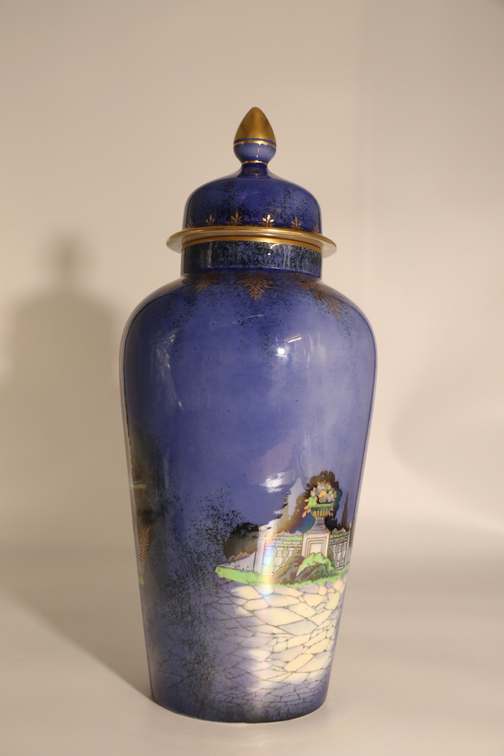 A cobalt blue ceramic lidded vase featuring a painted architectural garden scene with stone arches and gold accents. View IV