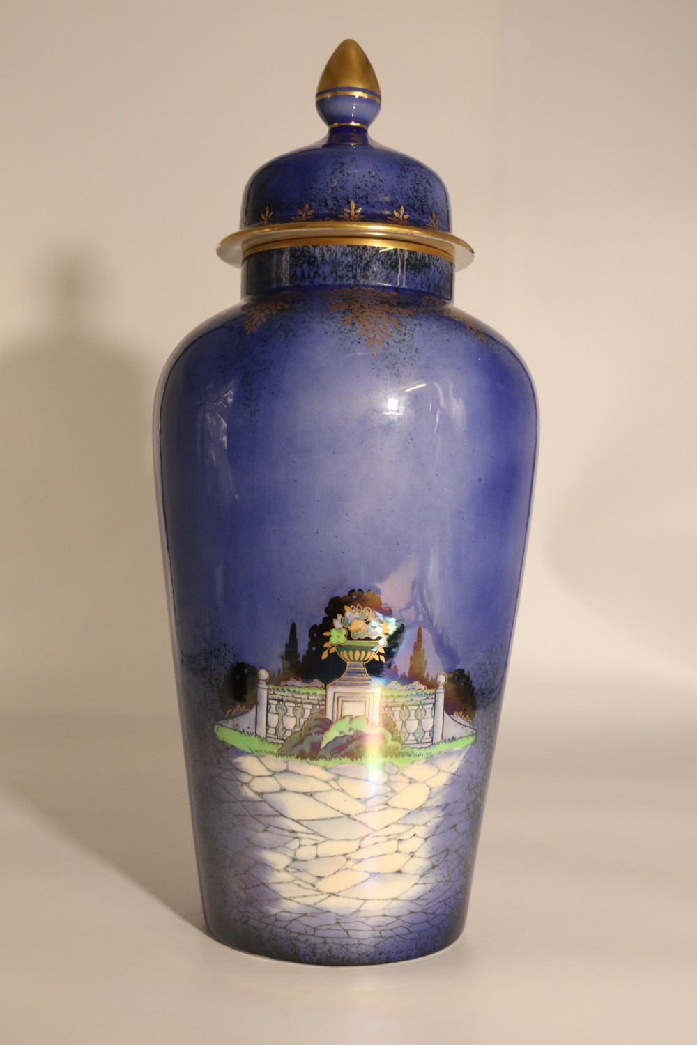 A cobalt blue ceramic lidded vase featuring a painted architectural garden scene with stone arches and gold accents. View V