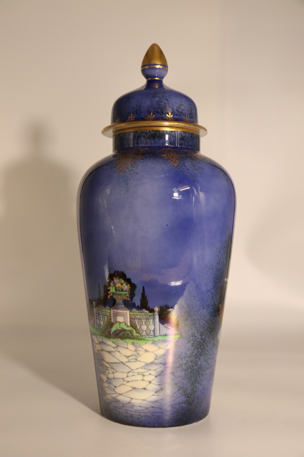 A cobalt blue ceramic lidded vase featuring a painted architectural garden scene with stone arches and gold accents. View VI