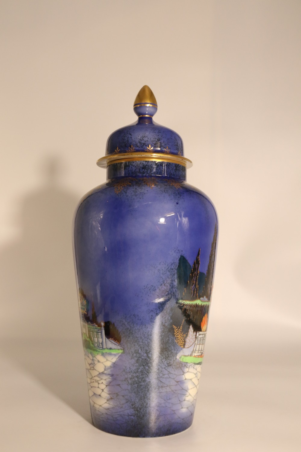A cobalt blue ceramic lidded vase featuring a painted architectural garden scene with stone arches and gold accents. View VII