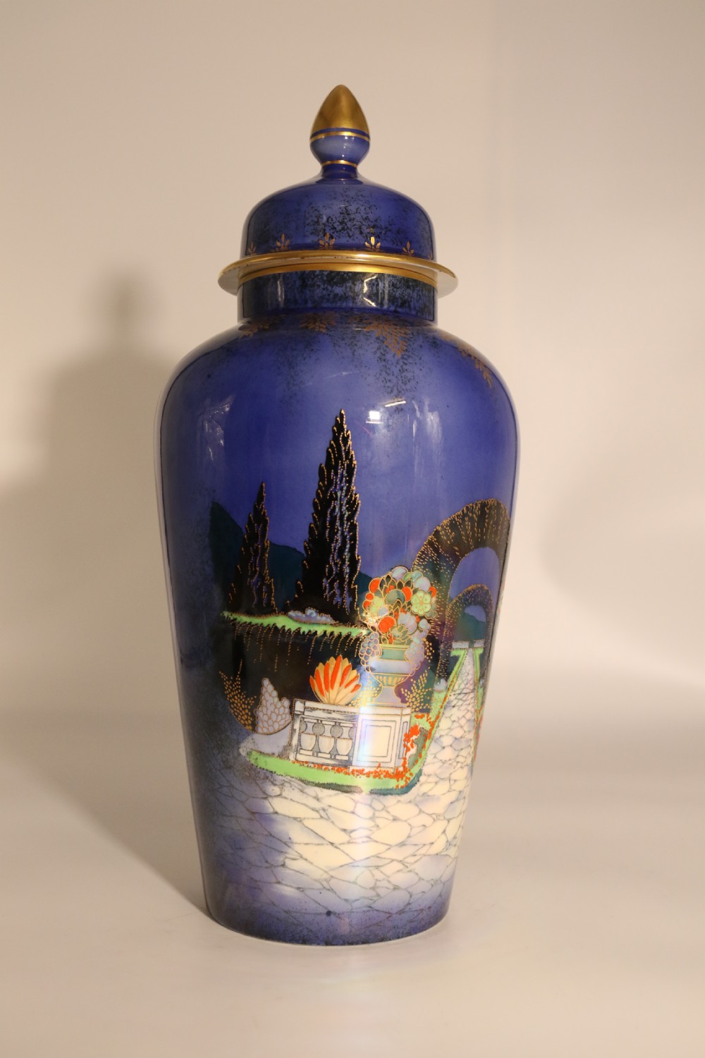 A cobalt blue ceramic lidded vase featuring a painted architectural garden scene with stone arches and gold accents. View IX