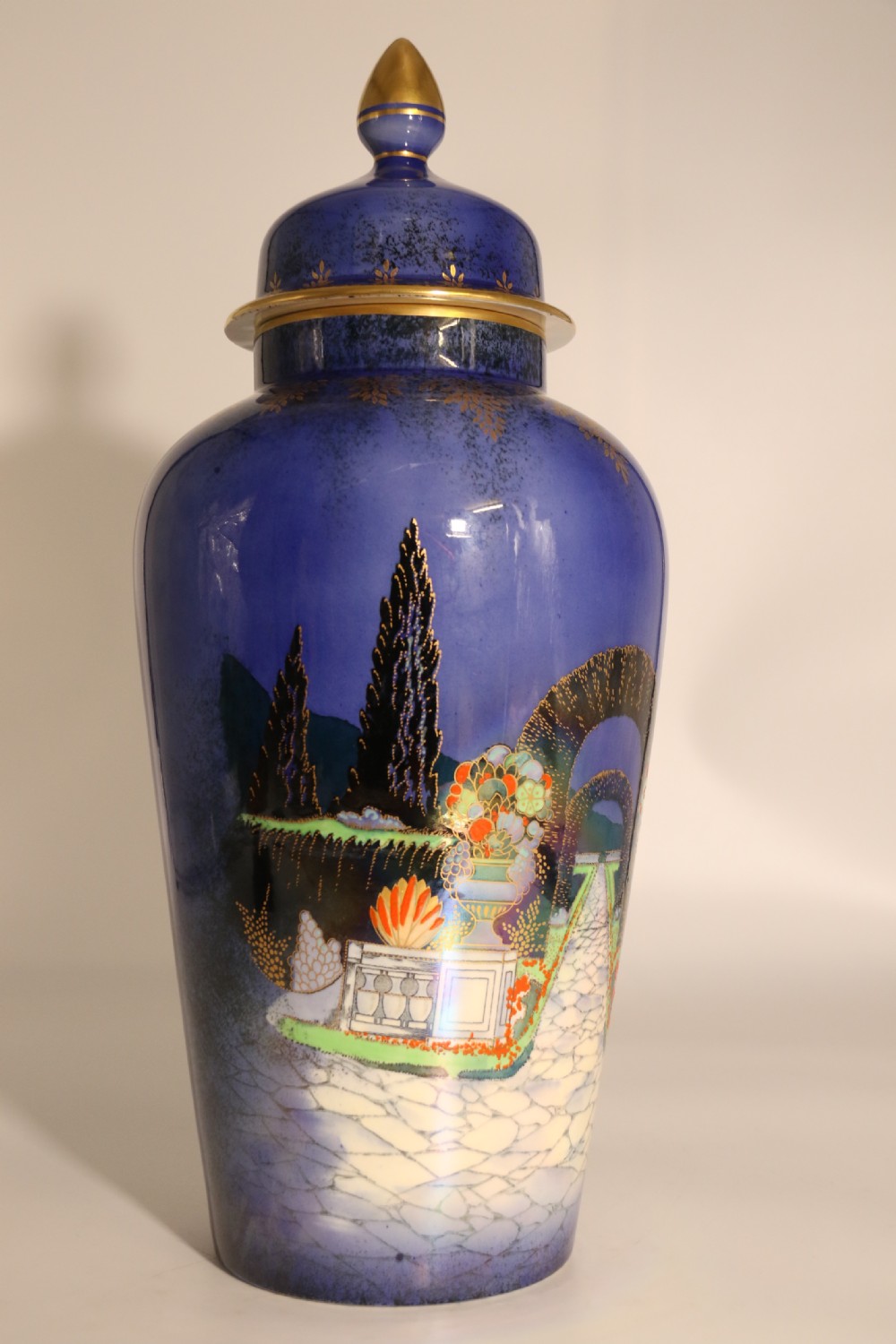A cobalt blue ceramic lidded vase featuring a painted architectural garden scene with stone arches and gold accents. View X