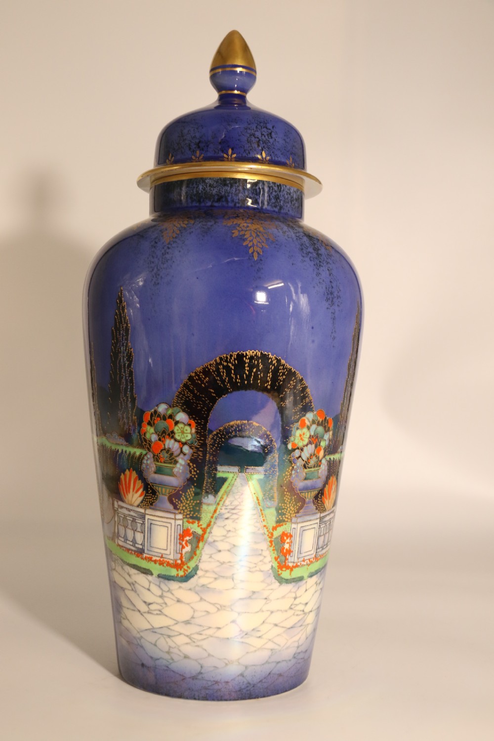 A cobalt blue ceramic lidded vase featuring a painted architectural garden scene with stone arches and gold accents. View XI