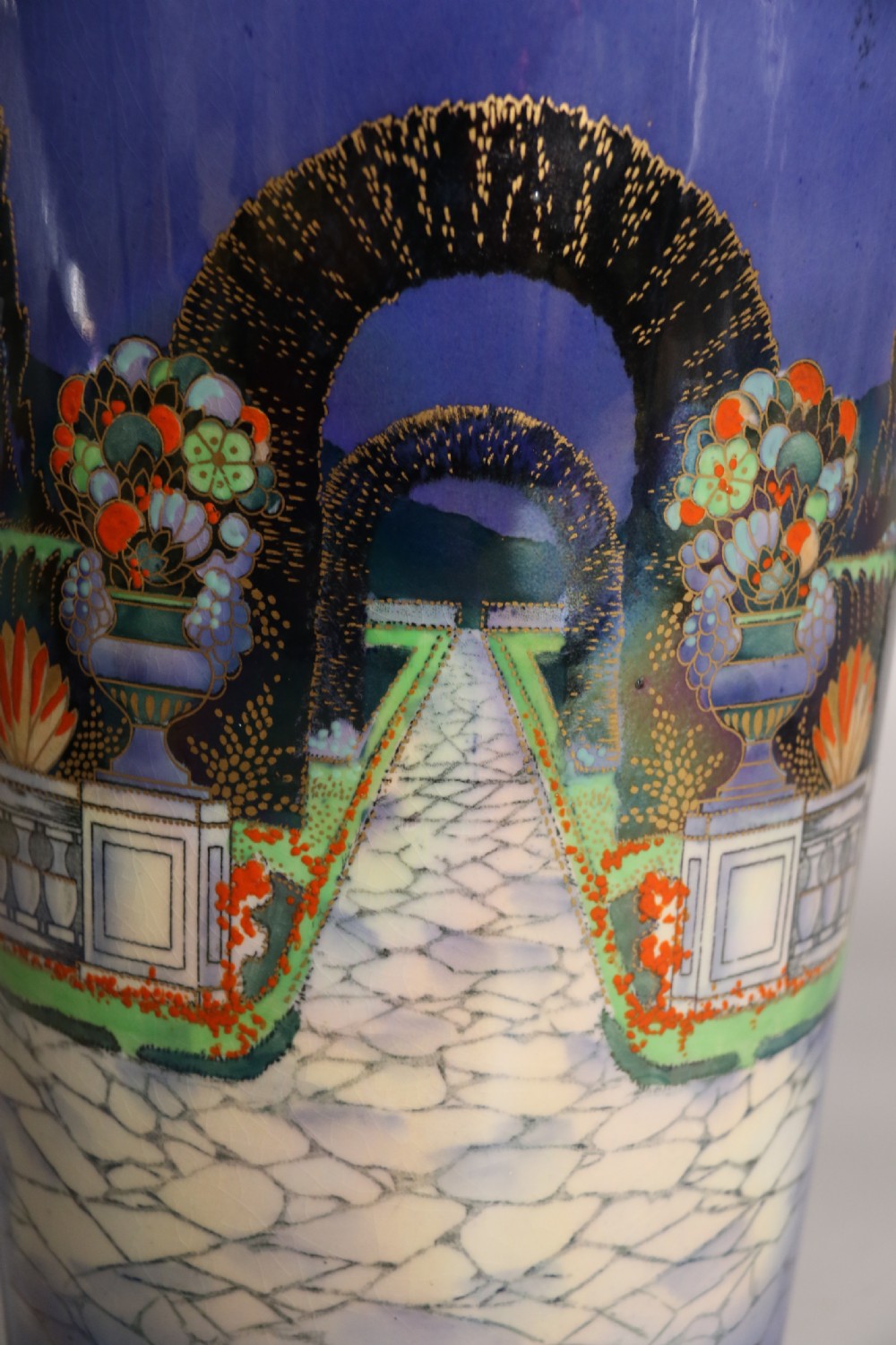 A cobalt blue ceramic lidded vase featuring a painted architectural garden scene with stone arches and gold accents. View XIV