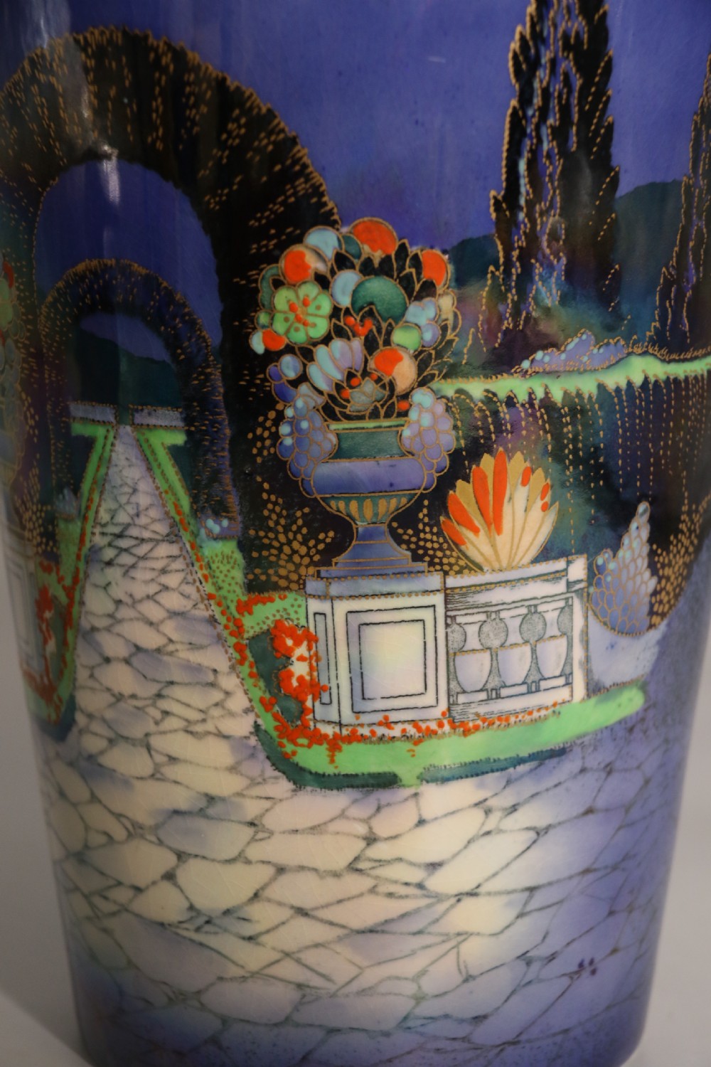 A cobalt blue ceramic lidded vase featuring a painted architectural garden scene with stone arches and gold accents. View XV