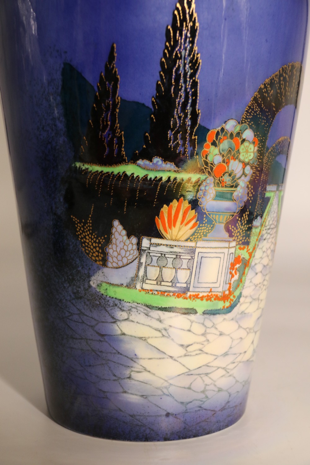 A cobalt blue ceramic lidded vase featuring a painted architectural garden scene with stone arches and gold accents. View XIX