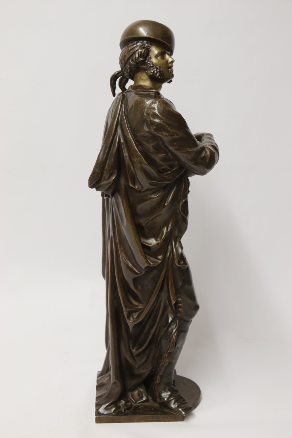 A patinated dark brown bronze sculpture depicting a standing male musician playing a mandolin while wearing a draped coat and headwear. View IV
