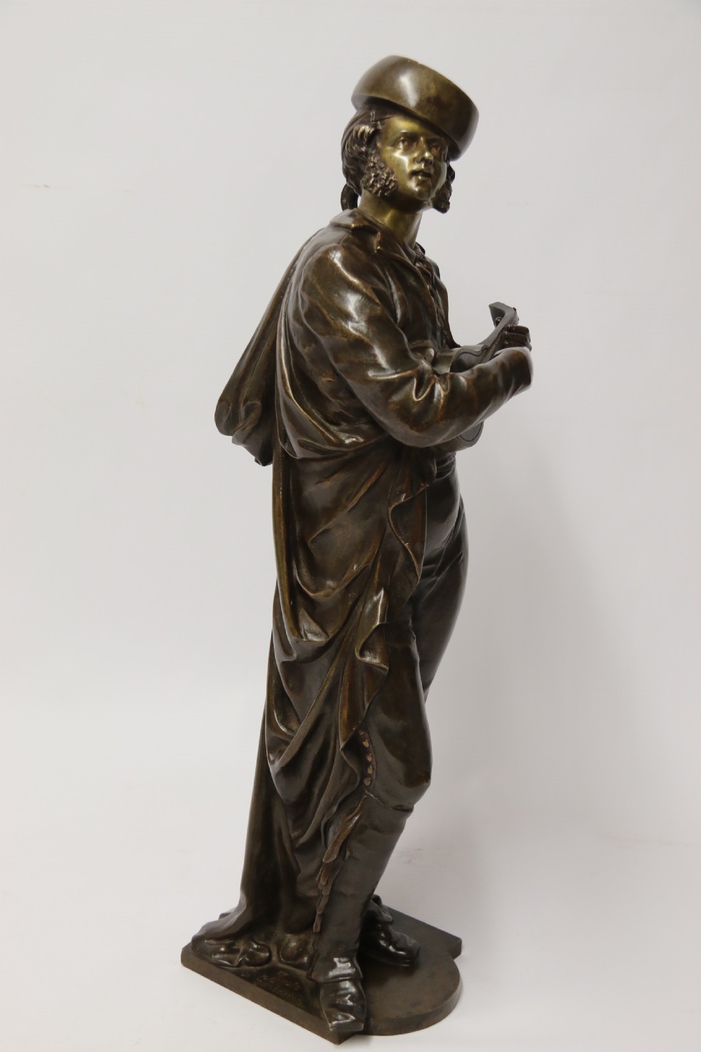 A patinated dark brown bronze sculpture depicting a standing male musician playing a mandolin while wearing a draped coat and headwear. View V