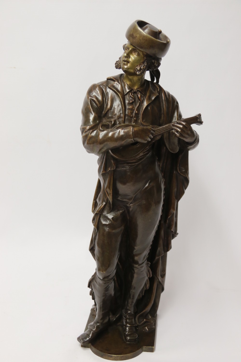 A patinated dark brown bronze sculpture depicting a standing male musician playing a mandolin while wearing a draped coat and headwear. View IX