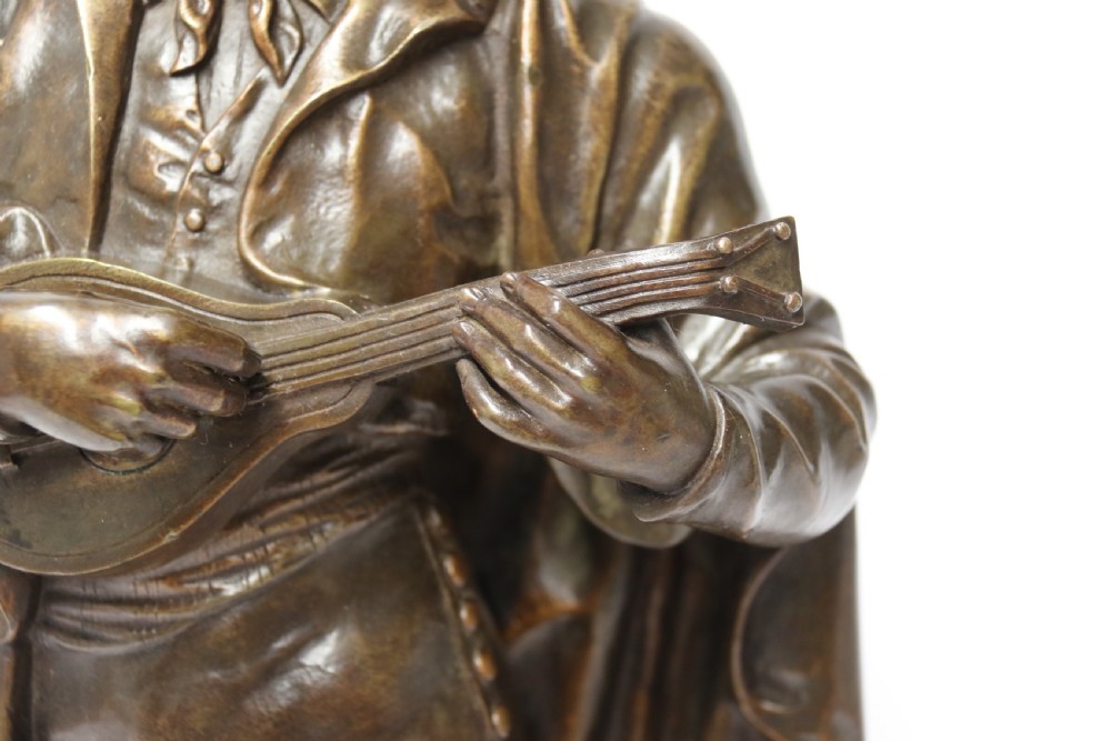A patinated dark brown bronze sculpture depicting a standing male musician playing a mandolin while wearing a draped coat and headwear. View XI
