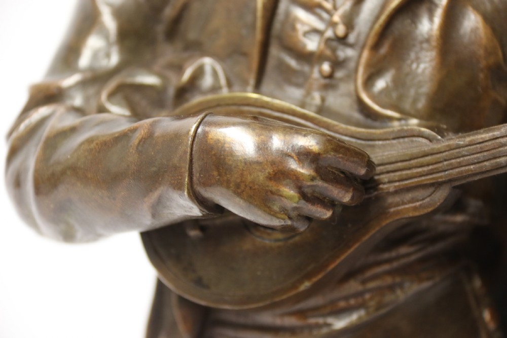 A patinated dark brown bronze sculpture depicting a standing male musician playing a mandolin while wearing a draped coat and headwear. View XII