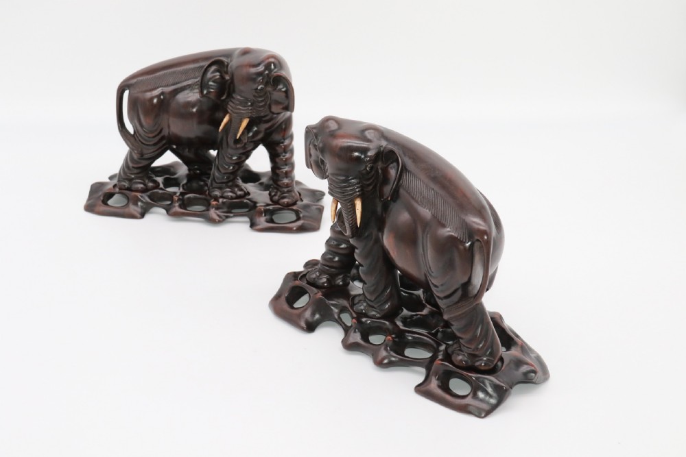 A pair of hand-carved dark hardwood elephant figures standing on pierced wooden bases with exposed tusks. View X