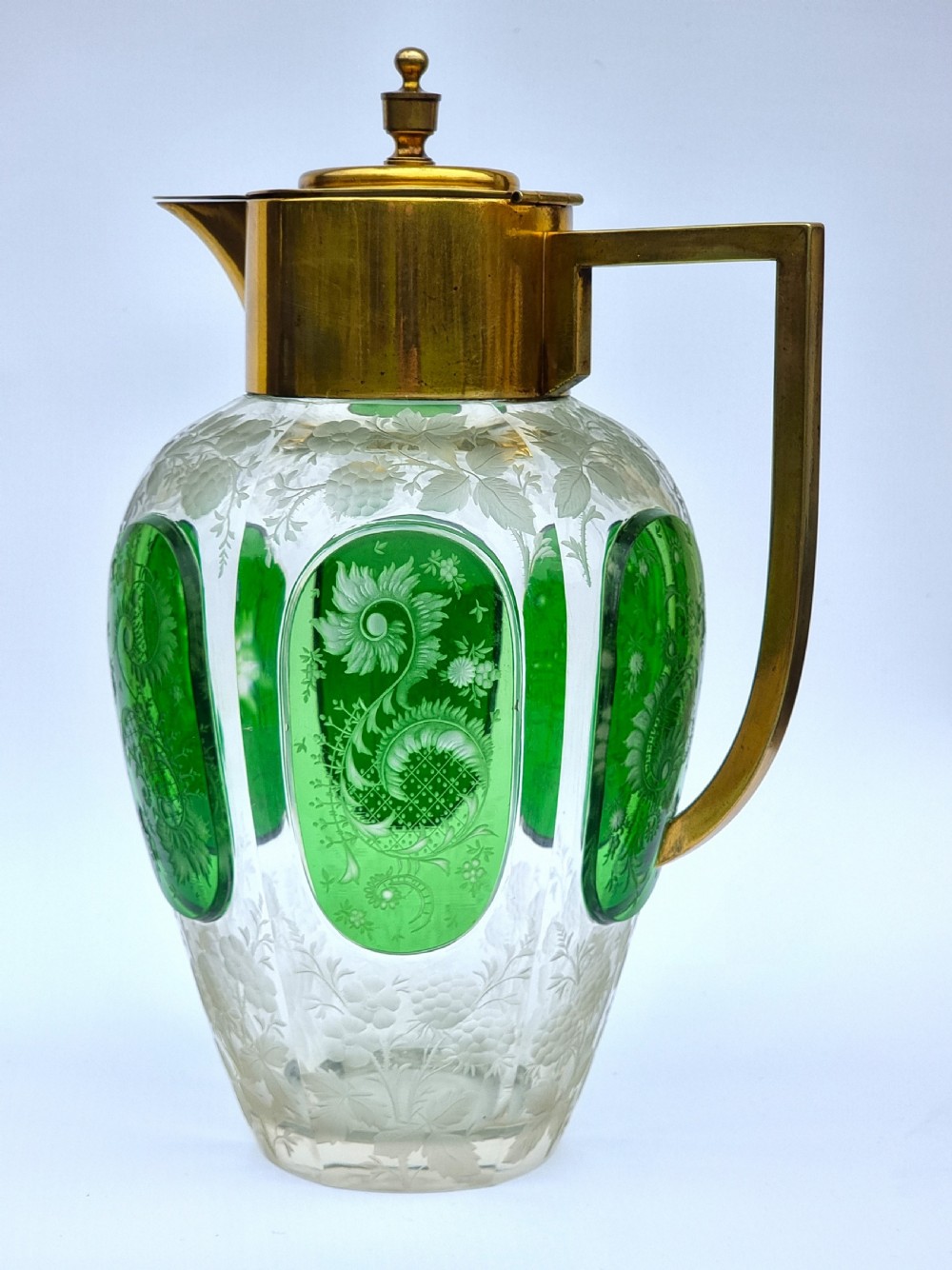 A clear glass claret jug featuring green overlay panels etched with floral motifs, topped with a metal collar, spout, lid, and geometric handle.