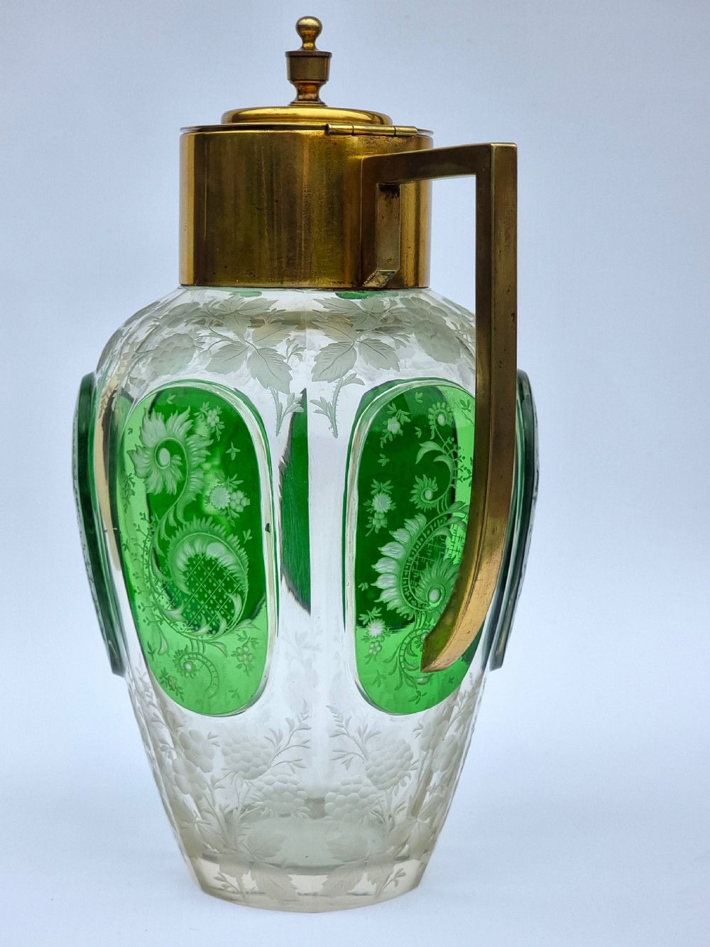 A clear glass claret jug featuring green overlay panels etched with floral motifs, topped with a metal collar, spout, lid, and geometric handle. View III