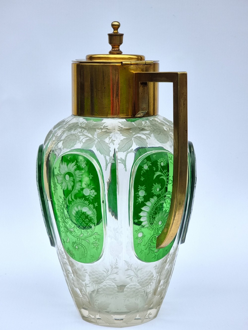 A clear glass claret jug featuring green overlay panels etched with floral motifs, topped with a metal collar, spout, lid, and geometric handle. View IV
