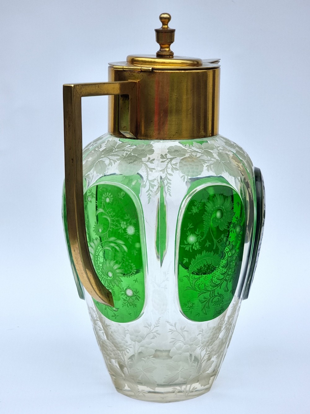 A clear glass claret jug featuring green overlay panels etched with floral motifs, topped with a metal collar, spout, lid, and geometric handle. View V