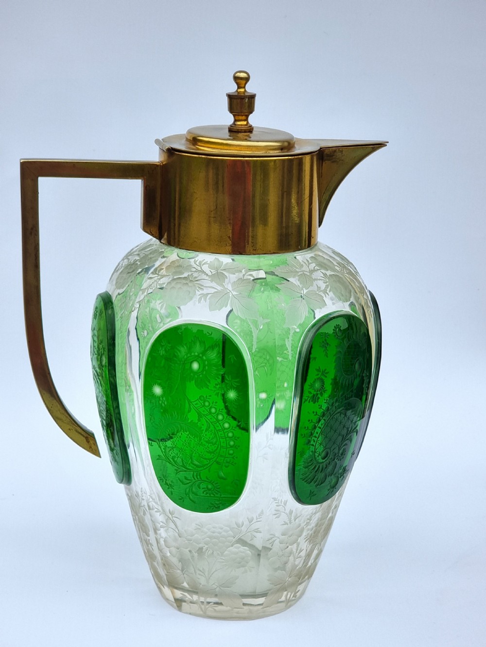 A clear glass claret jug featuring green overlay panels etched with floral motifs, topped with a metal collar, spout, lid, and geometric handle. View VI