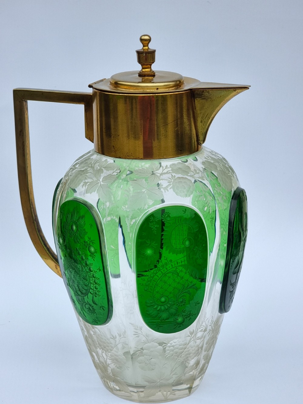 A clear glass claret jug featuring green overlay panels etched with floral motifs, topped with a metal collar, spout, lid, and geometric handle. View VII