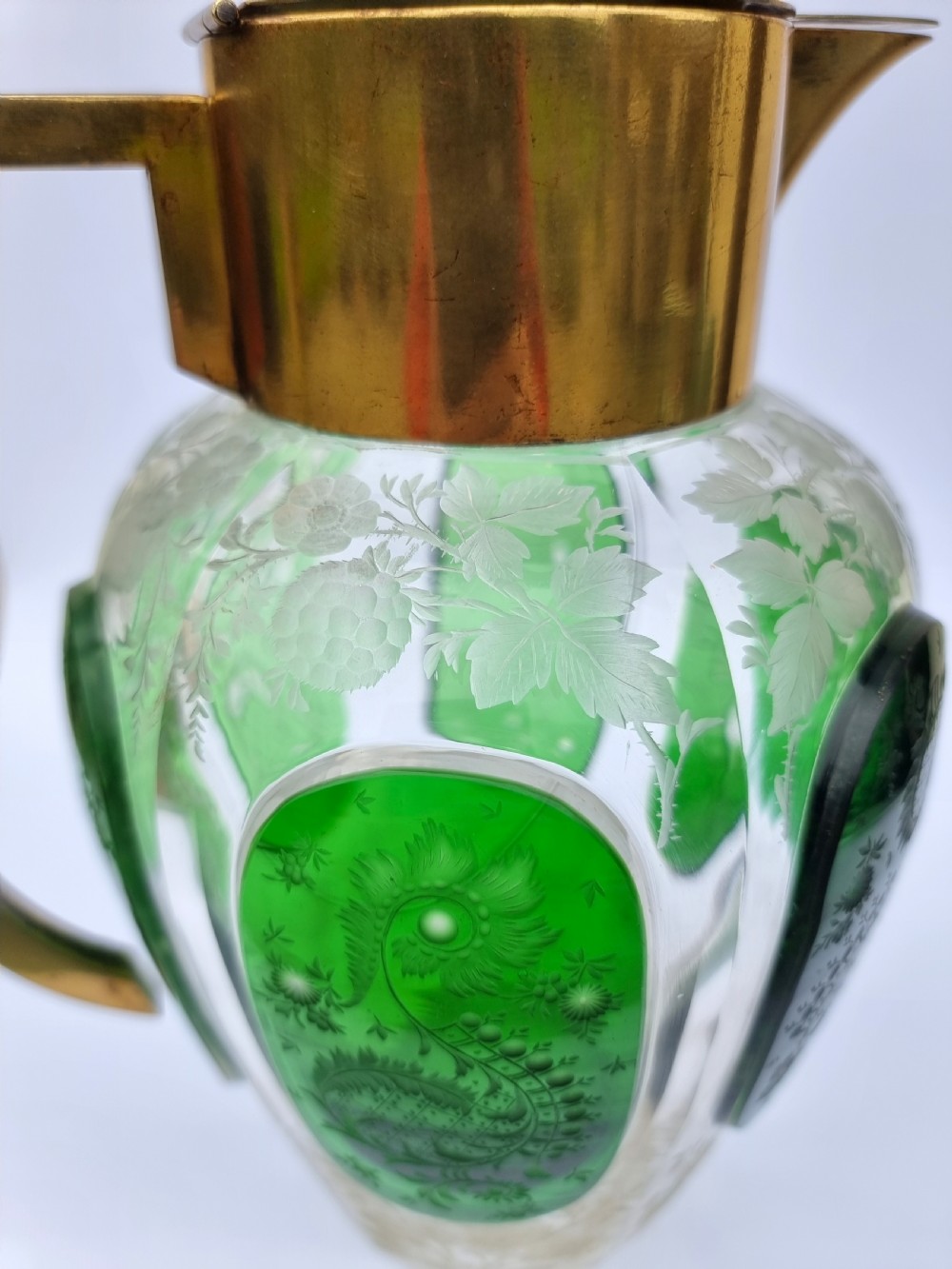 A clear glass claret jug featuring green overlay panels etched with floral motifs, topped with a metal collar, spout, lid, and geometric handle. View XIV
