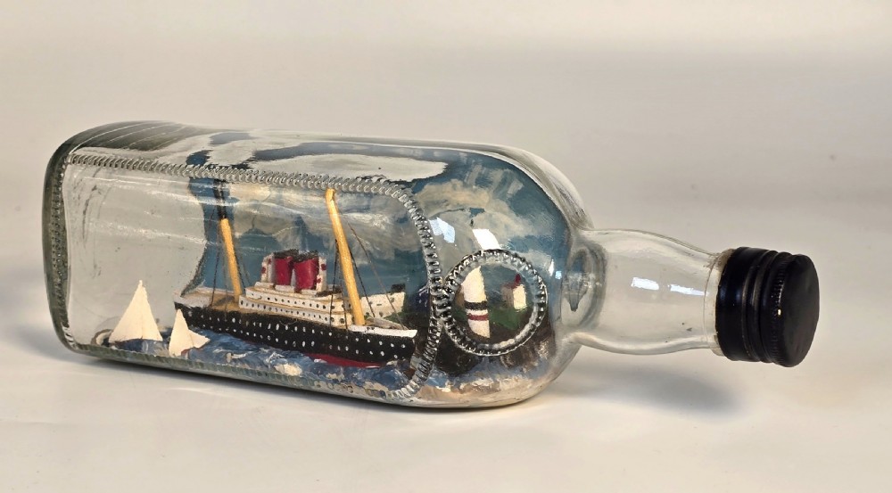 A horizontal clear glass bottle containing a detailed 1930s folk art diorama of a black, white, and red steamship on painted blue water with a lighthouse backdrop. View II