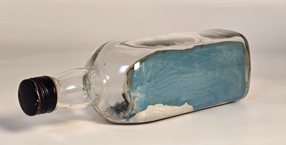 A horizontal clear glass bottle containing a detailed 1930s folk art diorama of a black, white, and red steamship on painted blue water with a lighthouse backdrop. View IV