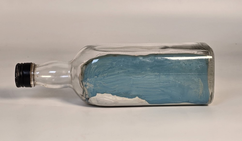 A horizontal clear glass bottle containing a detailed 1930s folk art diorama of a black, white, and red steamship on painted blue water with a lighthouse backdrop. View V