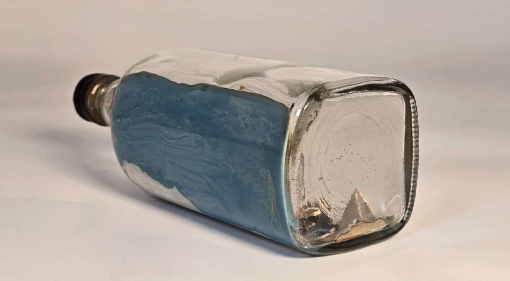 A horizontal clear glass bottle containing a detailed 1930s folk art diorama of a black, white, and red steamship on painted blue water with a lighthouse backdrop. View VII