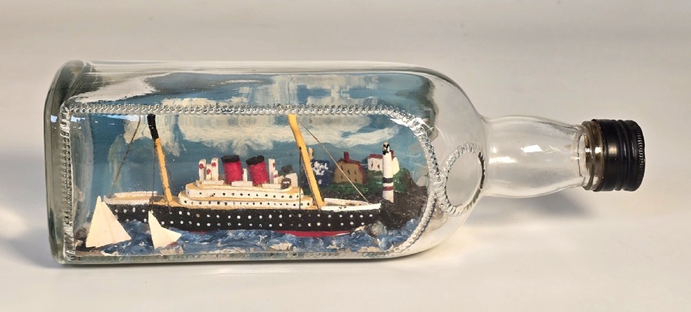 A horizontal clear glass bottle containing a detailed 1930s folk art diorama of a black, white, and red steamship on painted blue water with a lighthouse backdrop. View VIII