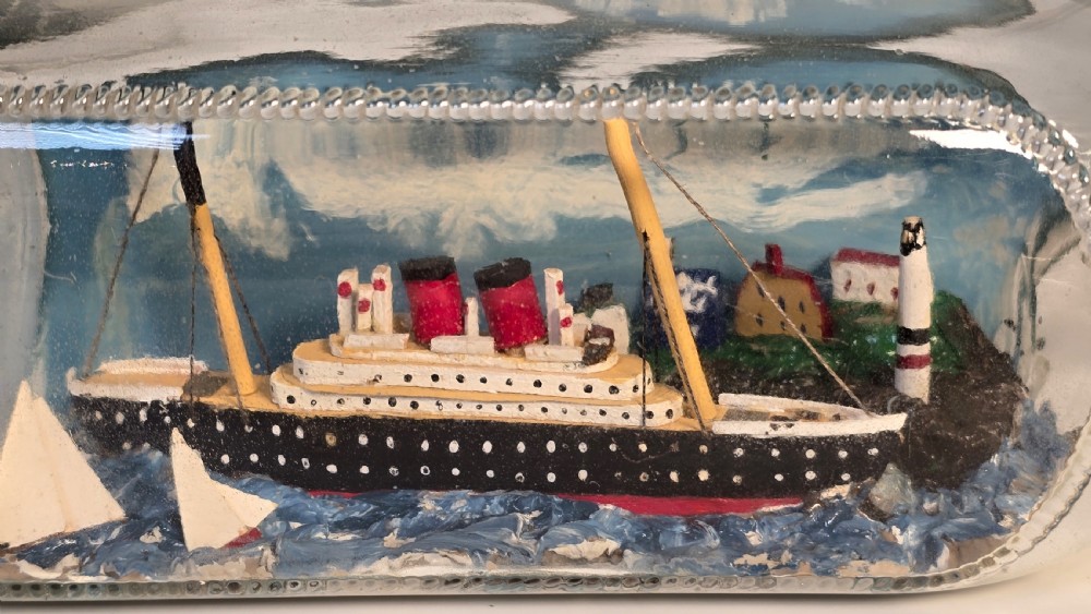 A horizontal clear glass bottle containing a detailed 1930s folk art diorama of a black, white, and red steamship on painted blue water with a lighthouse backdrop. View IX