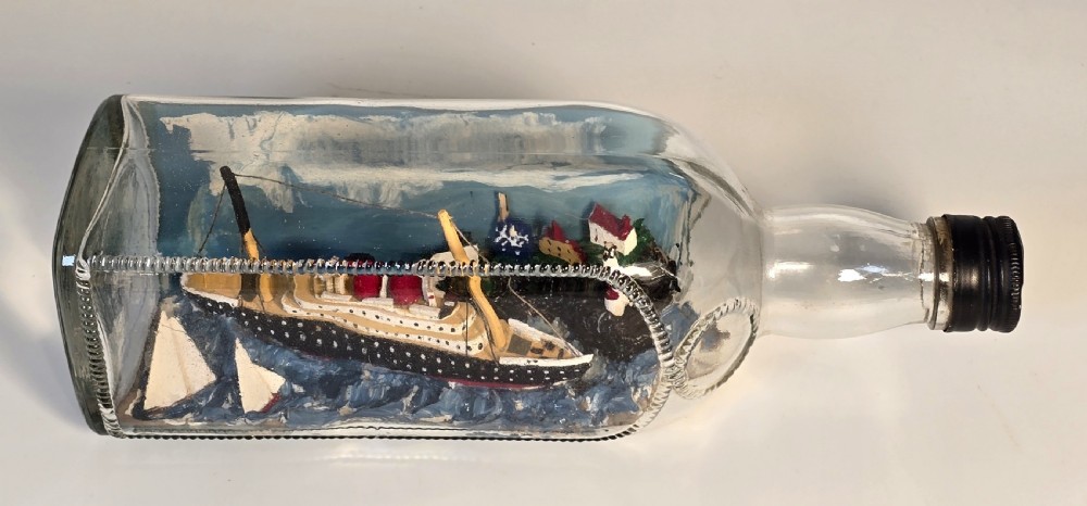 A horizontal clear glass bottle containing a detailed 1930s folk art diorama of a black, white, and red steamship on painted blue water with a lighthouse backdrop. View X