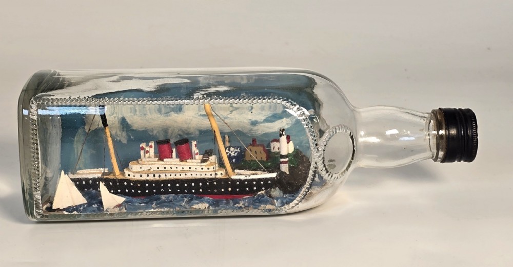 A horizontal clear glass bottle containing a detailed 1930s folk art diorama of a black, white, and red steamship on painted blue water with a lighthouse backdrop. View XI