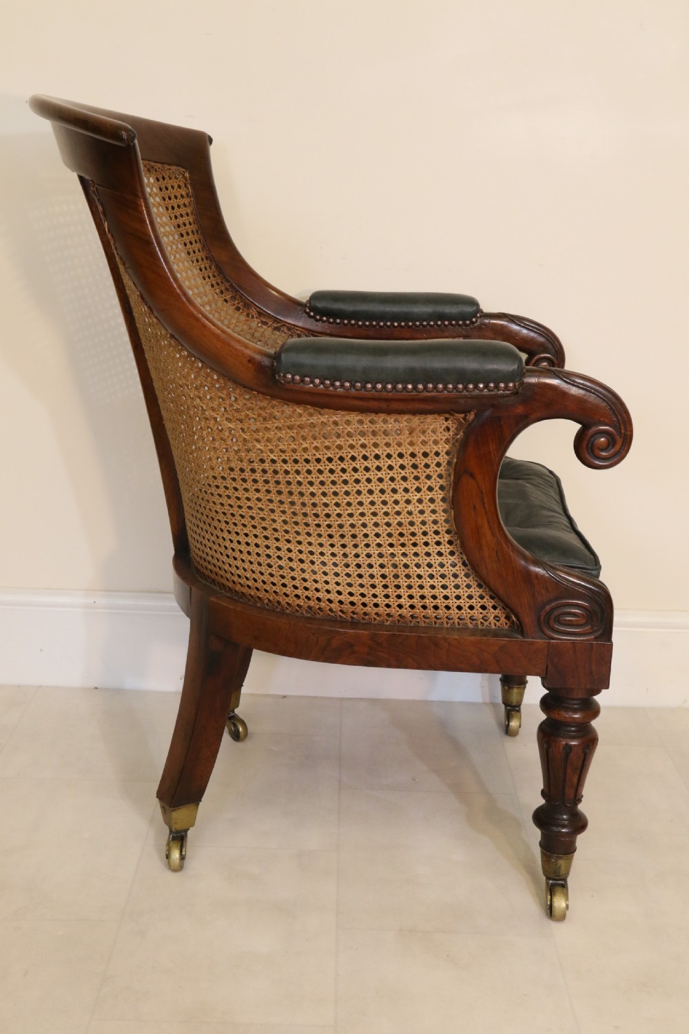 A pair of Regency rosewood bergere library armchairs with hand-woven cane panels, deep green leather cushions, scrolled arms, and turned legs on brass castors. View V